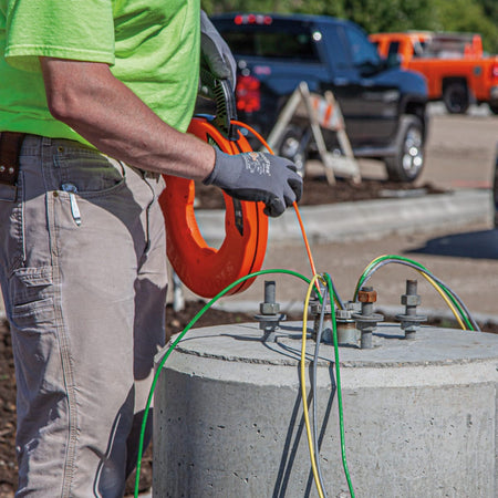 An outdoor application shot showing a contractor using the Klein Tools fiberglass fish tape to pull heavy-duty underground electrical cables through a concrete light pole base on a commercial jobsite.