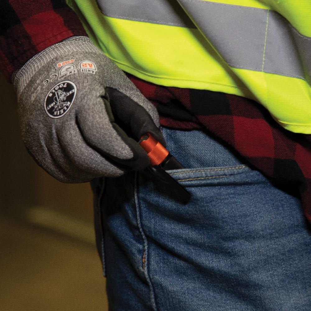 An application shot showing a worker wearing a safety glove and high-visibility vest placing the Klein Tools 56411 LED pocket light into a jeans pocket, demonstrating the secure hold of the dual-direction stainless steel clip.