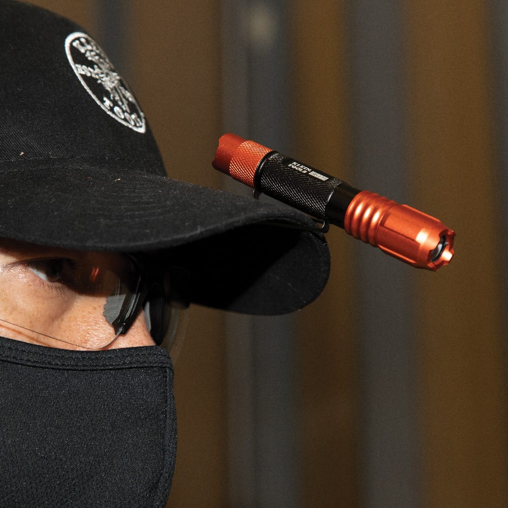 A close-up view of a professional wearing a black cap and safety glasses, with the rechargeable flashlight clipped to the bill of the hat to provide hands-free illumination for focused jobsite tasks.
