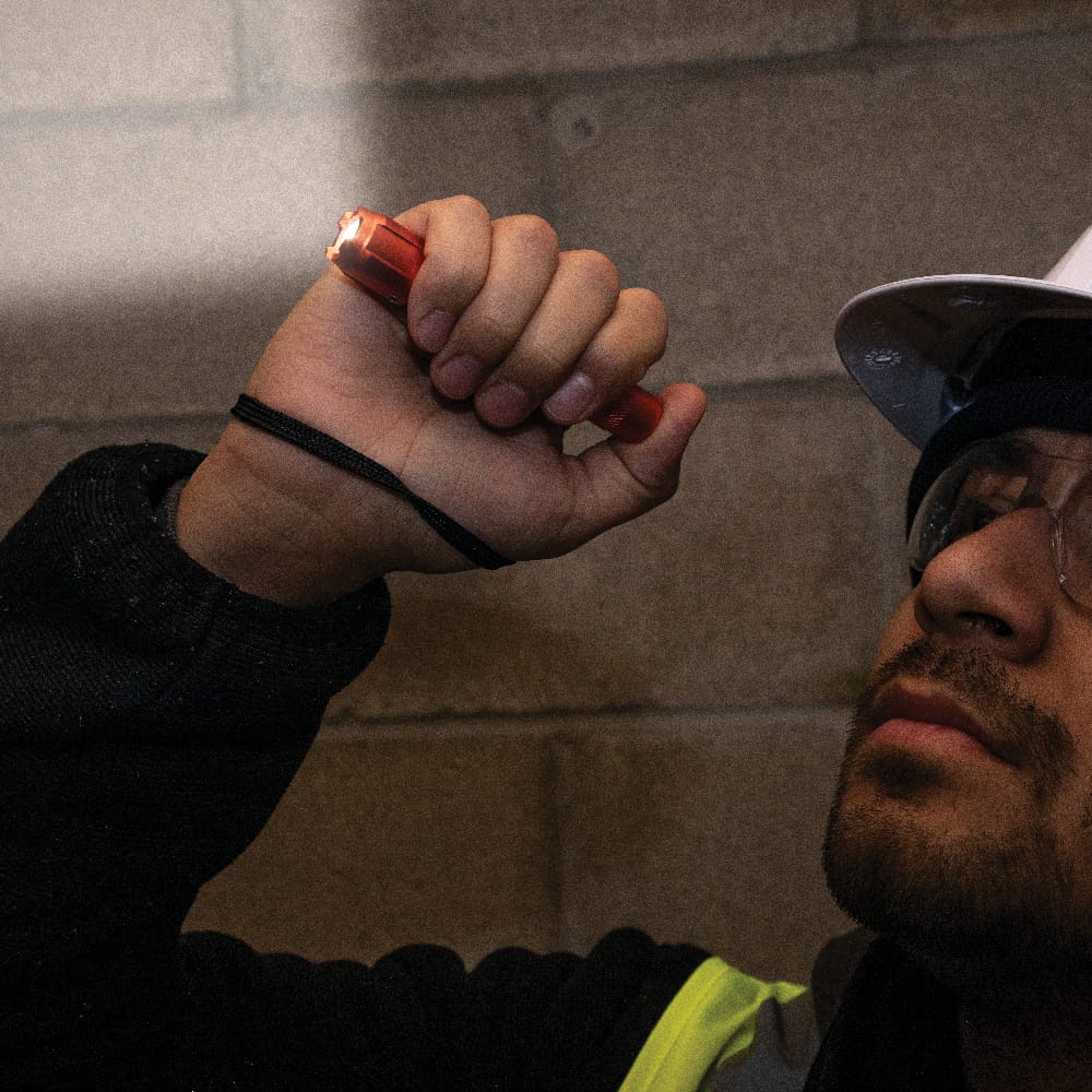 An application shot of a professional tradesperson in a hard hat and safety glasses using the rechargeable light to inspect a dark concrete block wall, demonstrating its compact size and high-intensity beam.