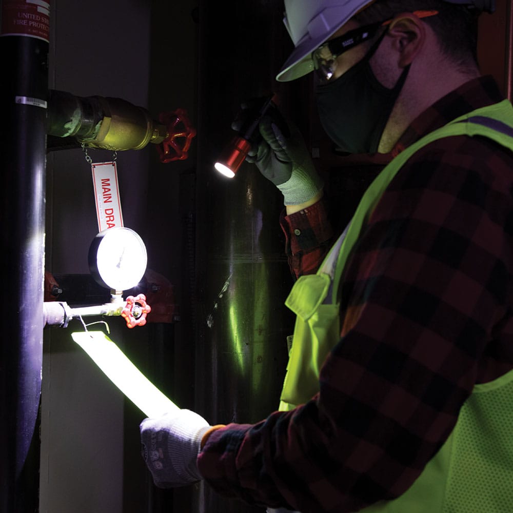 Professional worker wearing a hardhat and safety gear using the bright 500 lumen Klein Tools 56412 rechargeable flashlight to inspect a pipe gauge in a dark industrial setting.