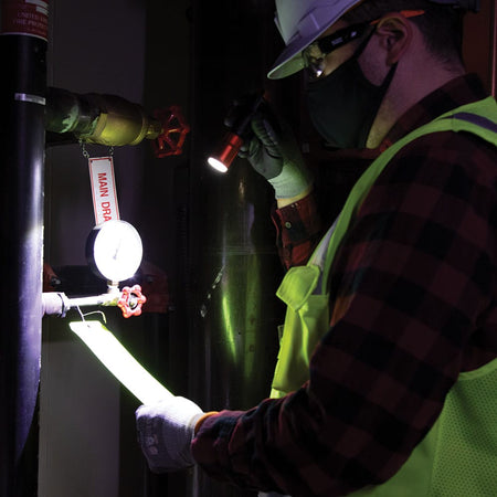 Professional worker wearing a hardhat and safety gear using the bright 500 lumen Klein Tools 56412 rechargeable flashlight to inspect a pipe gauge in a dark industrial setting.