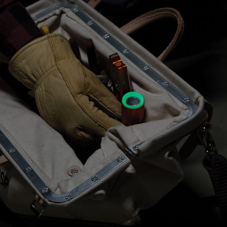 Gloved hand reaching into a dark tool bag to retrieve the Klein Tools 56412 IP66 water and dust proof flashlight, demonstrating how the bright green glow ring locator bezel makes it easy to find in low light.