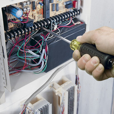 An action photograph showing a professional using the Klein Tools 601-6 3/16-inch cabinet tip screwdriver to tighten a screw on a complex electrical terminal block inside a control panel.