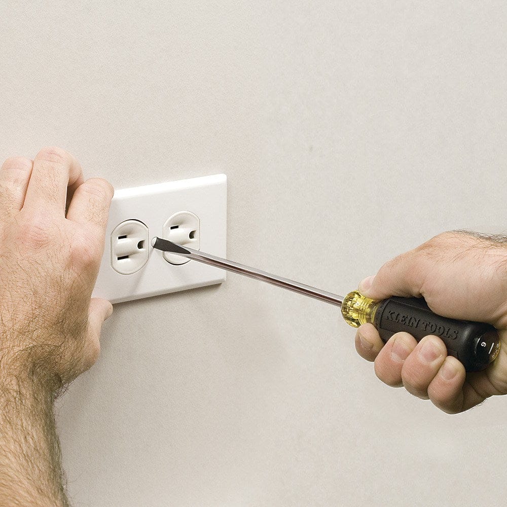 A close-up action shot of the Klein Tools 601-6 cabinet tip screwdriver being used to install a white duplex electrical outlet cover plate, demonstrating the precision of the narrow 3/16-inch tip.