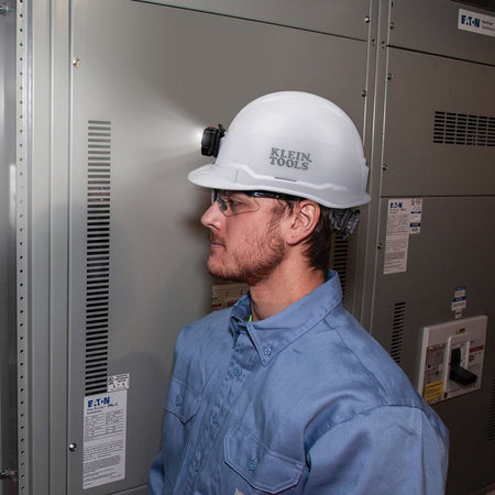 An application photo of a professional electrician wearing the Klein Tools white cap-style hard hat with the 300-lumen rechargeable LED headlamp illuminated while inspecting an electrical panel, demonstrating the non-vented Class E helmet's utility in specialized work environments.