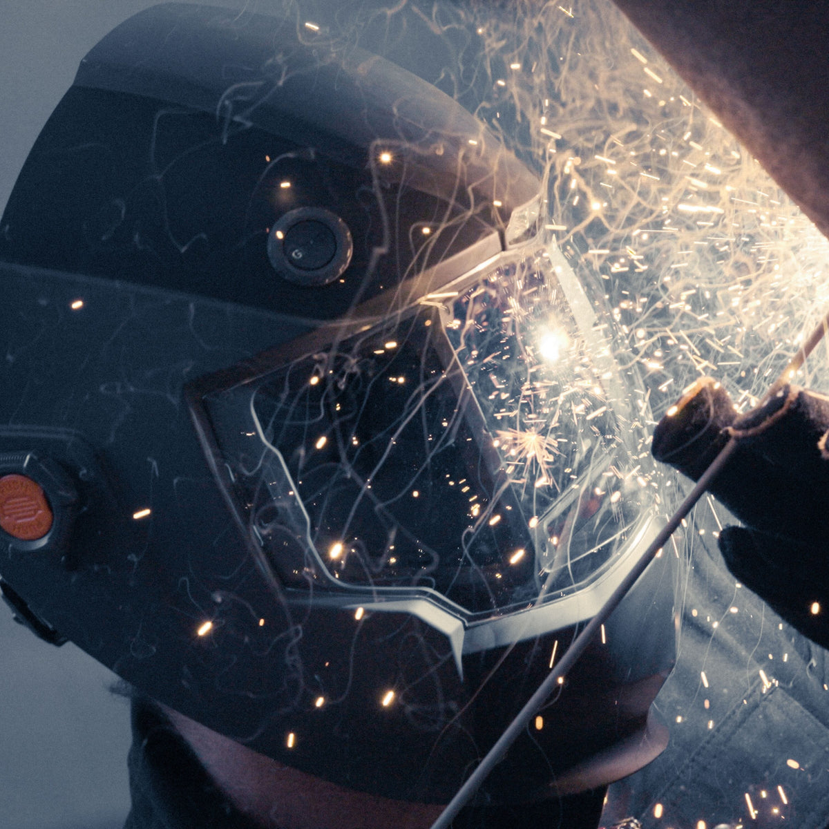 An action lifestyle shot of a professional technician wearing the Klein Tools 60141 helmet. The integrated LED headlamp is turned on, illuminating a metal workpiece in a dark environment to demonstrate the auto-work light feature.