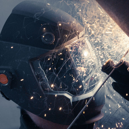 An action lifestyle shot of a professional technician wearing the Klein Tools 60141 helmet. The integrated LED headlamp is turned on, illuminating a metal workpiece in a dark environment to demonstrate the auto-work light feature.