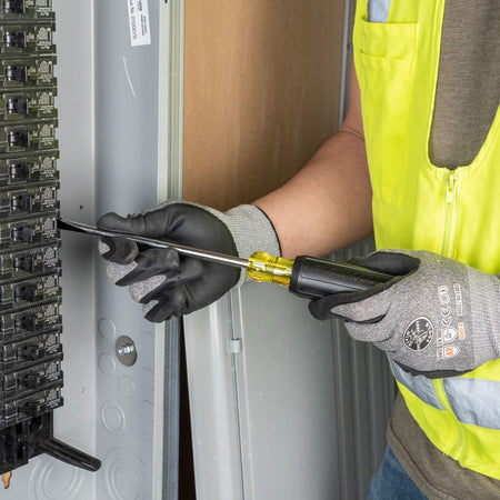 An electrician wearing protective safety gloves using the flat keystone tip of the Klein Tools 602-7DD demolition screwdriver to carefully align components inside an open grey metal electrical panel box.