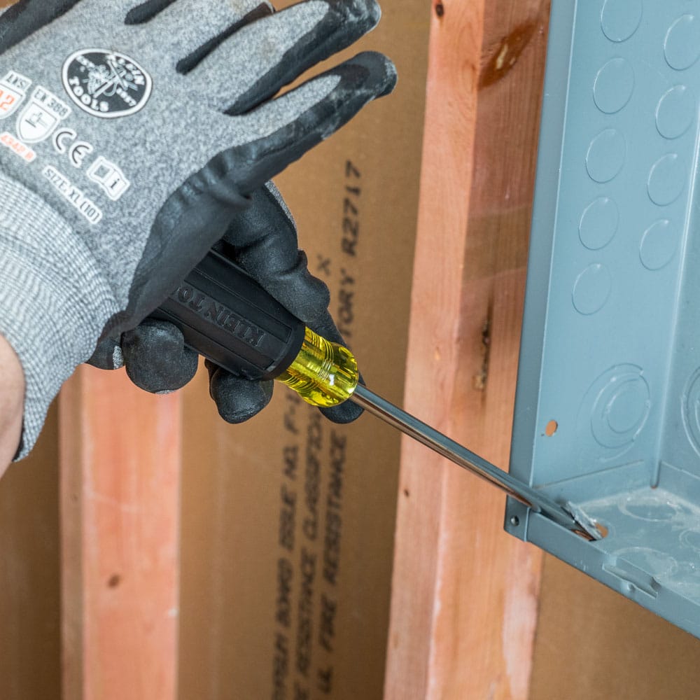 An active jobsite view of a worker utilizing the Klein Tools 602-7DD demolition screwdriver as a high-leverage prying hand tool to breakout a metal knock-out slug from a steel junction box against exposed wood framing.