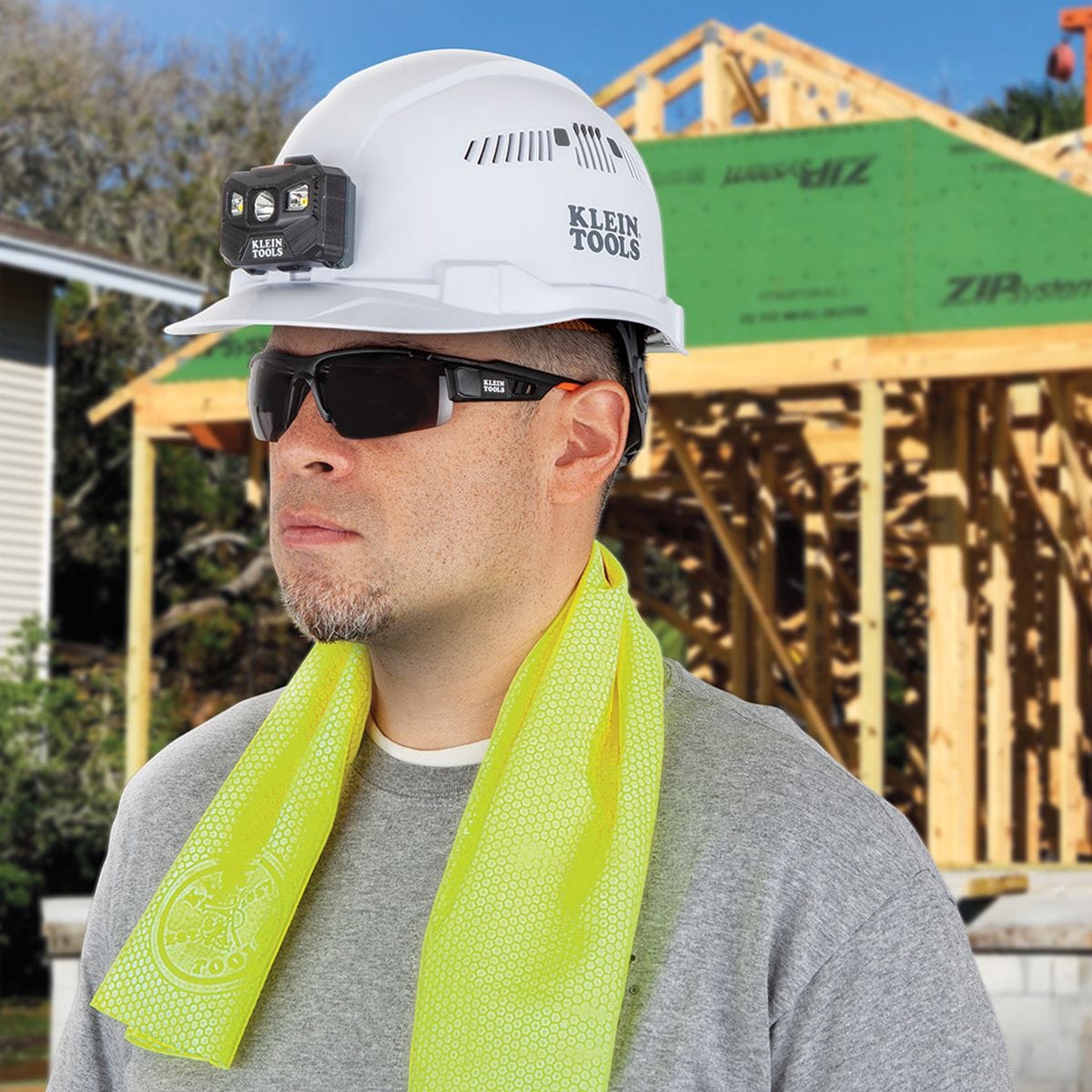 An outdoor jobsite lifestyle application photo of a male construction worker wearing a white safety hard hat with an attached headlamp, dark safety glasses, and a grey shirt, with a high-visibility yellow Klein Tools evaporative cooling towel draped around his neck in front of a residential house framing background.