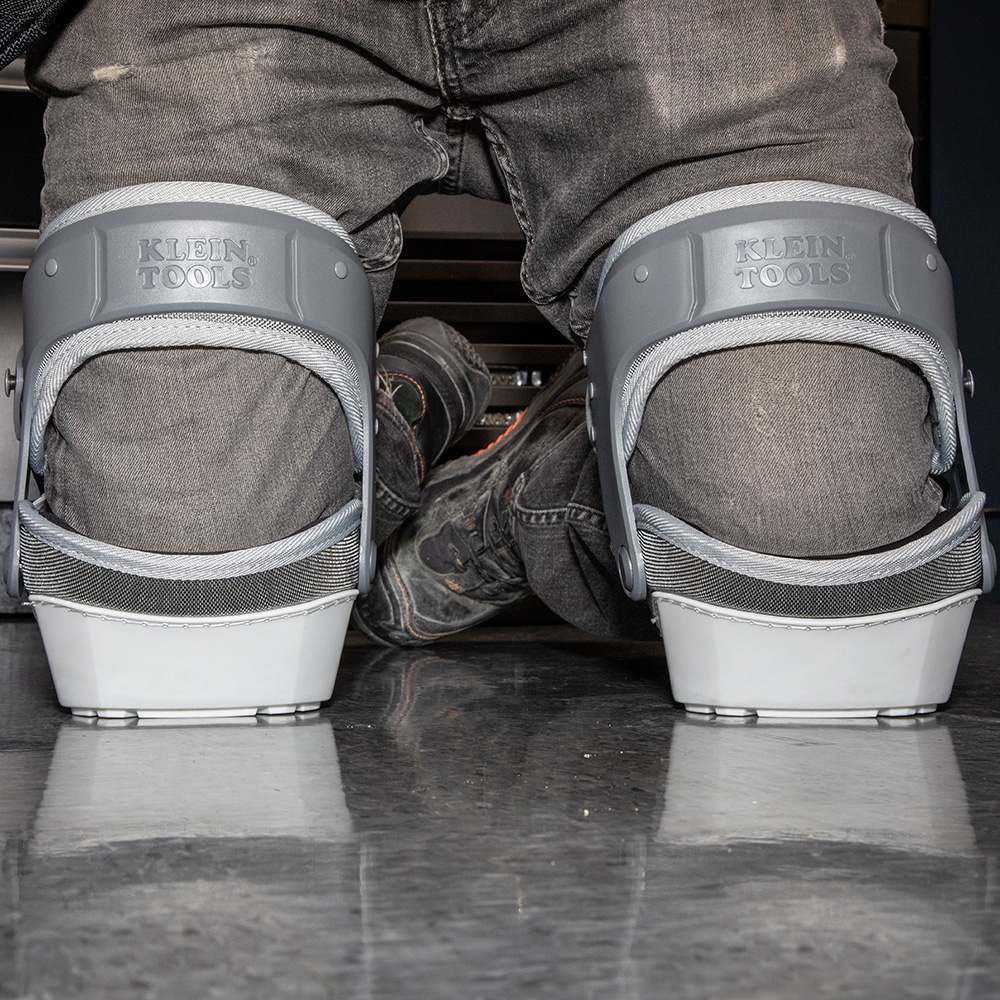 A rear view of a worker kneeling on a polished floor, showcasing the back of the Klein Tools stabilizer knee pads. The image highlights the secure fit of the gray hinged straps and the stability provided by the flat-bottomed shells.