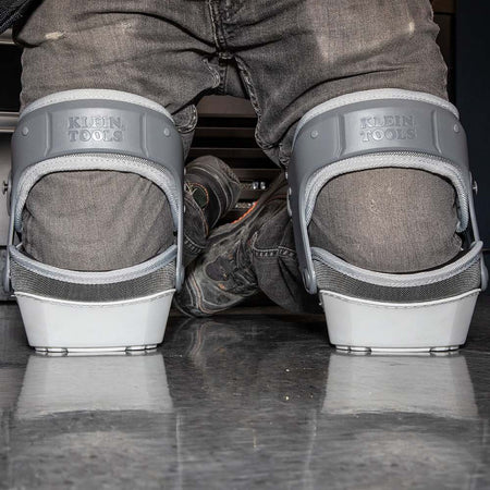 A rear view of a worker kneeling on a polished floor, showcasing the back of the Klein Tools stabilizer knee pads. The image highlights the secure fit of the gray hinged straps and the stability provided by the flat-bottomed shells.