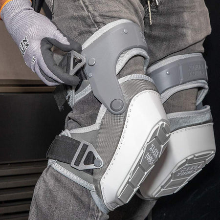 A close-up shot of a gloved hand adjusting the quick-fasten buckle on the gray hinged thigh strap. This demonstrates the ease of achieving a secure and customizable fit for optimal knee protection.