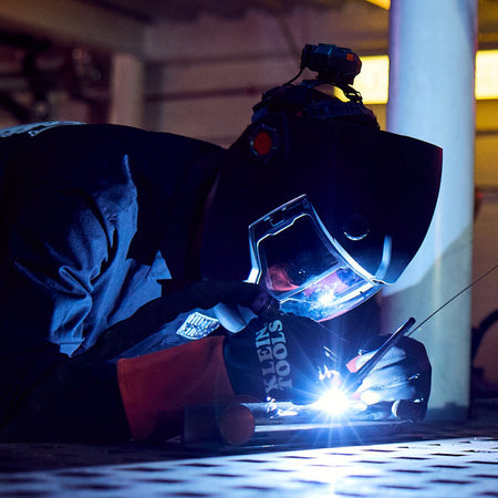 A high-contrast action shot of a professional welder in a dark environment. The technician is wearing the Klein Tools 60912 TIG gloves and a Phoenix Series welding helmet, demonstrating the precise torch control and dexterity provided by the full-grain cowhide construction.