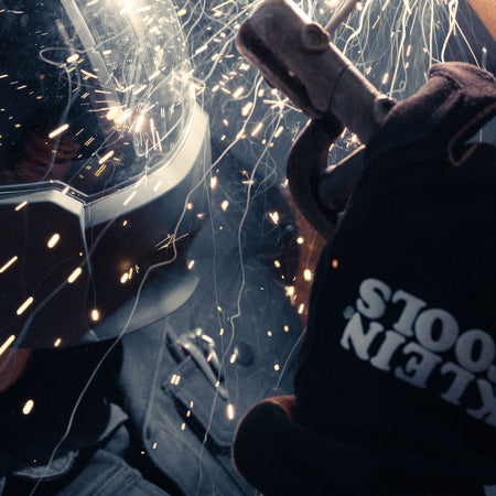 A high-action lifestyle shot of a technician performing a heavy-duty weld. The professional is wearing the Klein Tools 60914 MIG gloves, which are shown resisting a shower of molten sparks and spatter, demonstrating the extreme thermal protection of the split cowhide leather.