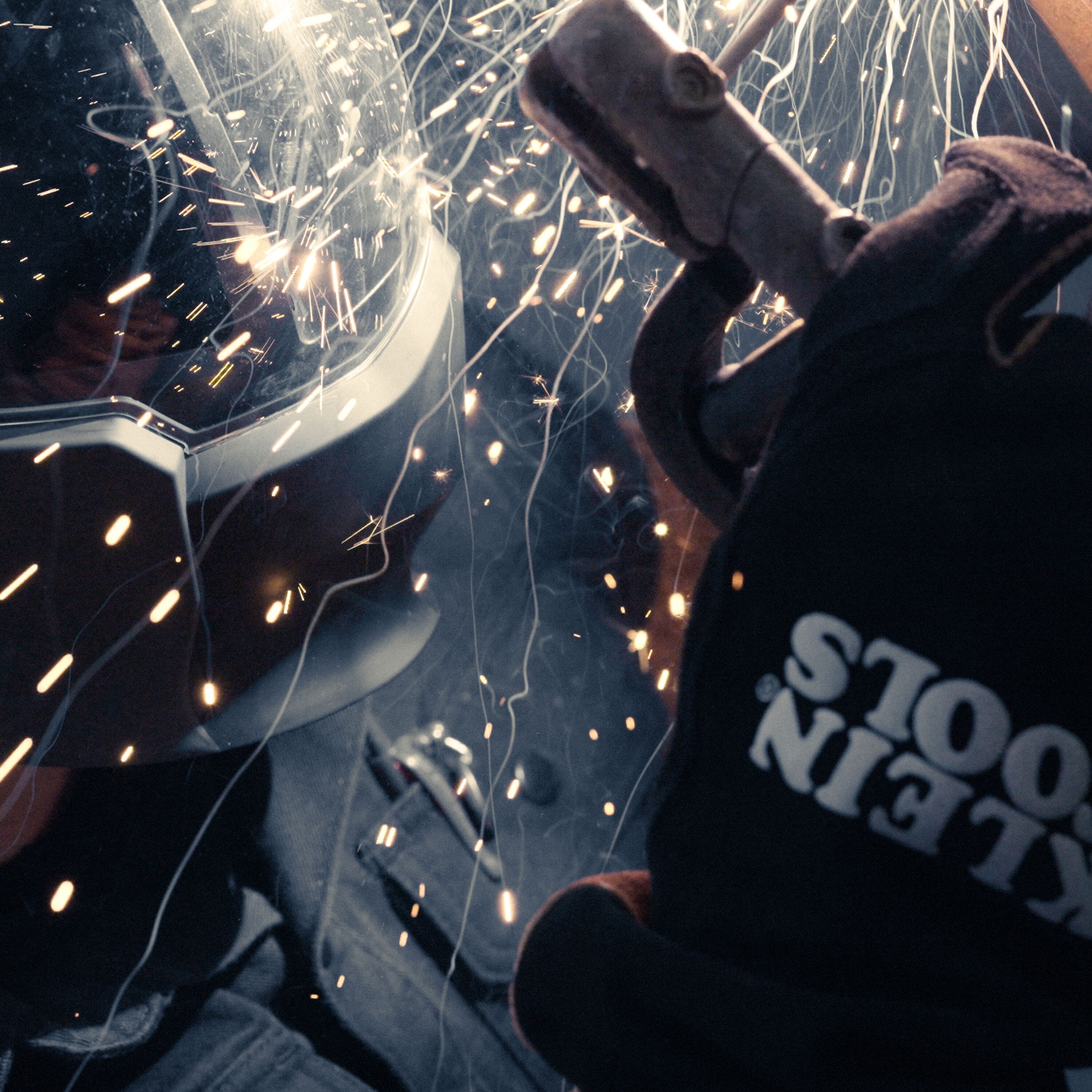A high-action lifestyle shot of a technician performing a heavy-duty weld. The professional is wearing the Klein Tools 60915 MIG gloves, which are shown resisting a shower of molten sparks and spatter, demonstrating the extreme thermal protection of the split cowhide leather.