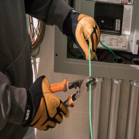 An action shot of a professional tradesperson wearing protective gloves and using the Klein Tools insulated cable cutter to perform a clean shear cut on a green communication cable near an electrical panel.