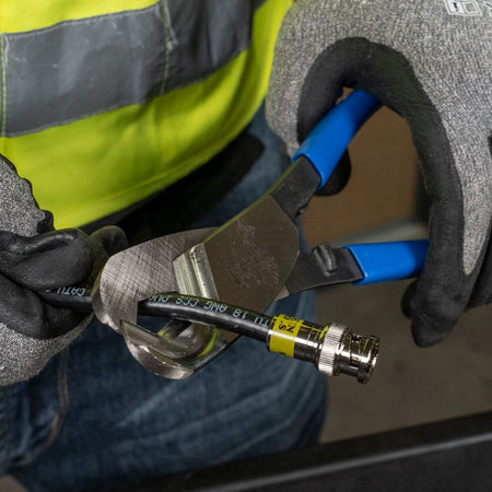 An action shot of a professional tradesperson using the Klein Tools coaxial cutter to perform a clean, one-handed shear cut on a black coaxial cable fitted with a BNC connector.