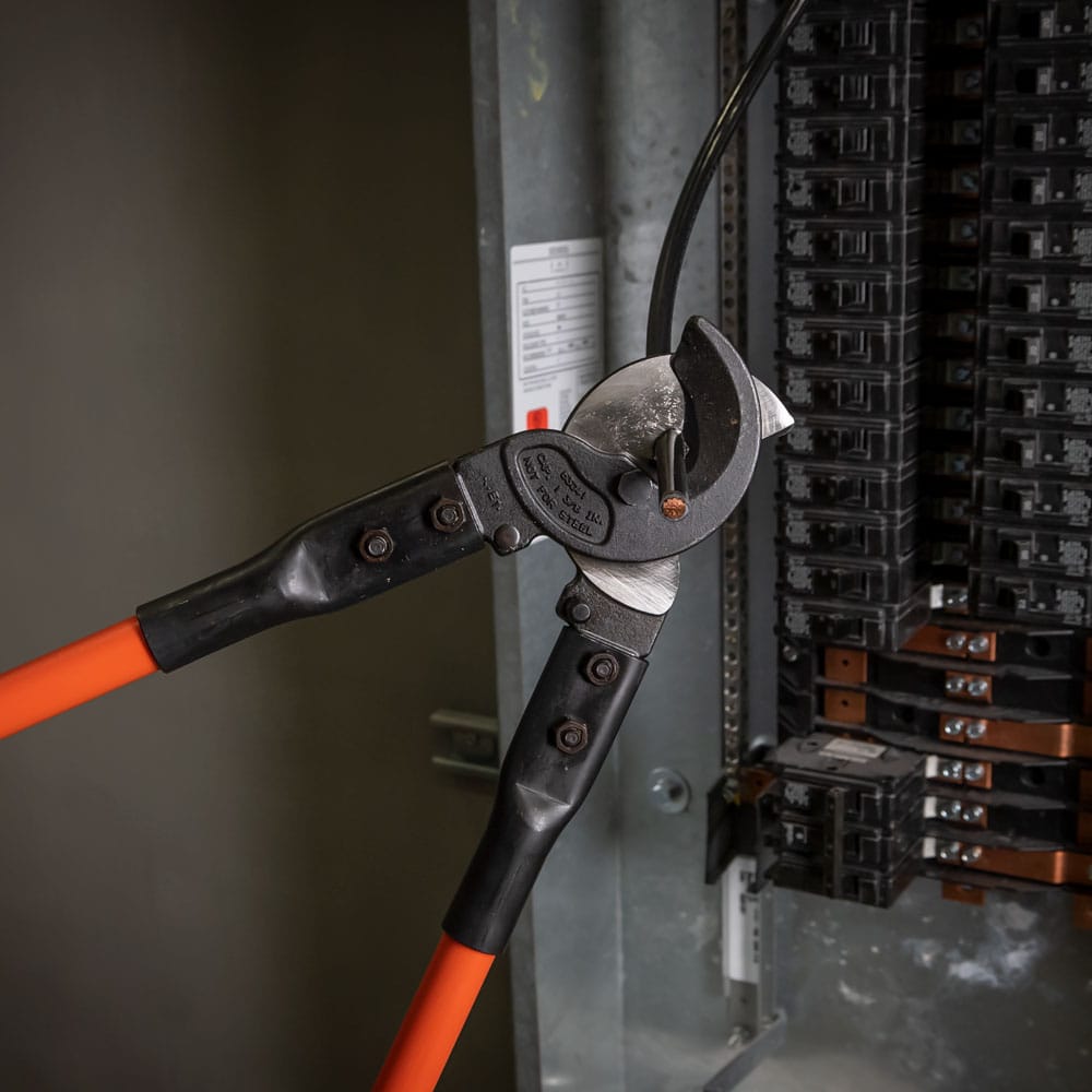 An action shot of the Klein Tools cable cutter being used to perform a clean shear cut on a heavy-duty copper conductor inside a commercial electrical service panel, highlighting its effectiveness in professional installations.