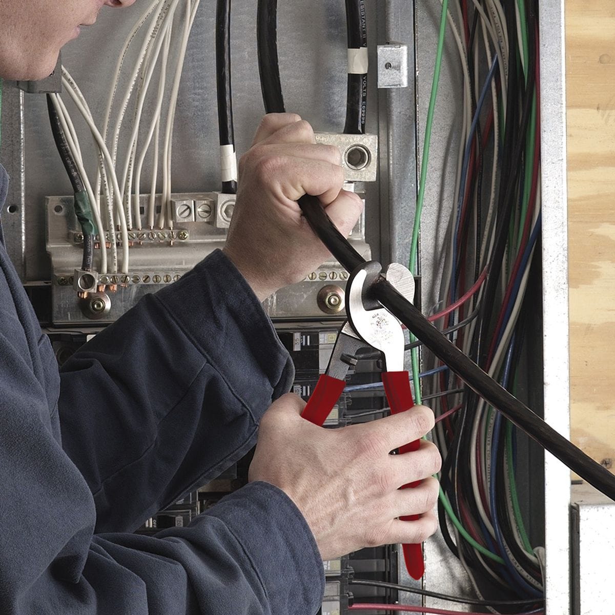 An action shot of an electrician using the 63050 cable cutter inside a commercial electrical panel to perform a clean, burr-free shear cut on a heavy-gauge black conductor, demonstrating its efficiency in confined spaces.