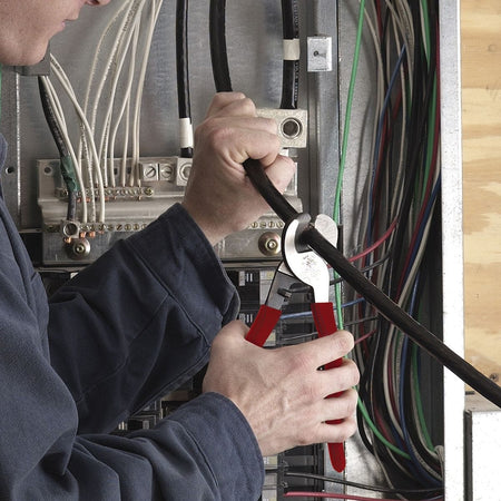 An action shot of an electrician using the 63050 cable cutter inside a commercial electrical panel to perform a clean, burr-free shear cut on a heavy-gauge black conductor, demonstrating its efficiency in confined spaces.