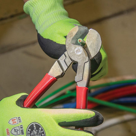 A close-up application photo of a professional tradesperson in green safety gloves using the 63225 high-leverage cable cutter to perform a clinical, burr-free shear cut on a small insulated conductor.