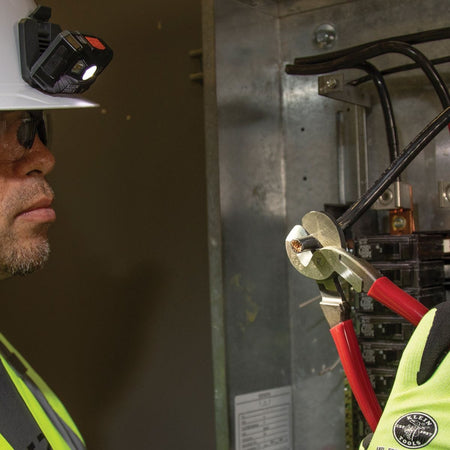 A jobsite action photo of a professional electrician in a hard hat and safety glasses utilizing the 63225 high-leverage cutter inside a commercial service panel to shear a heavy-gauge black insulated cable.