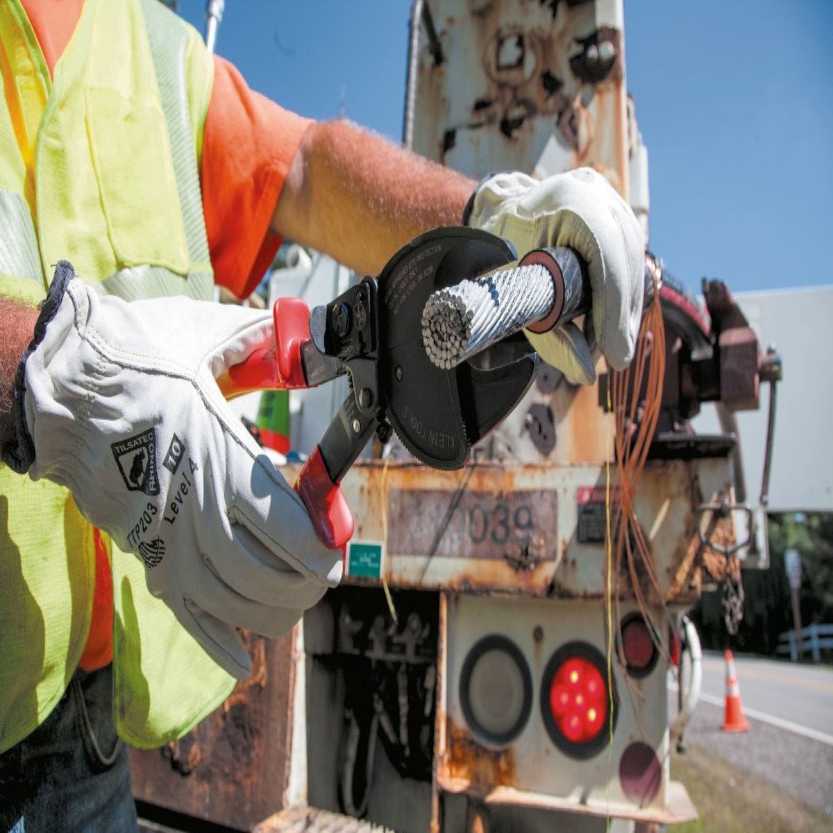 An outdoor action shot of a utility worker wearing safety gear and gloves while using the Klein Tools 63711 open-jaw ratcheting cutter to perform a clean, high-leverage cut on a large-diameter aluminum cable, demonstrating its professional-grade performance in field conditions.