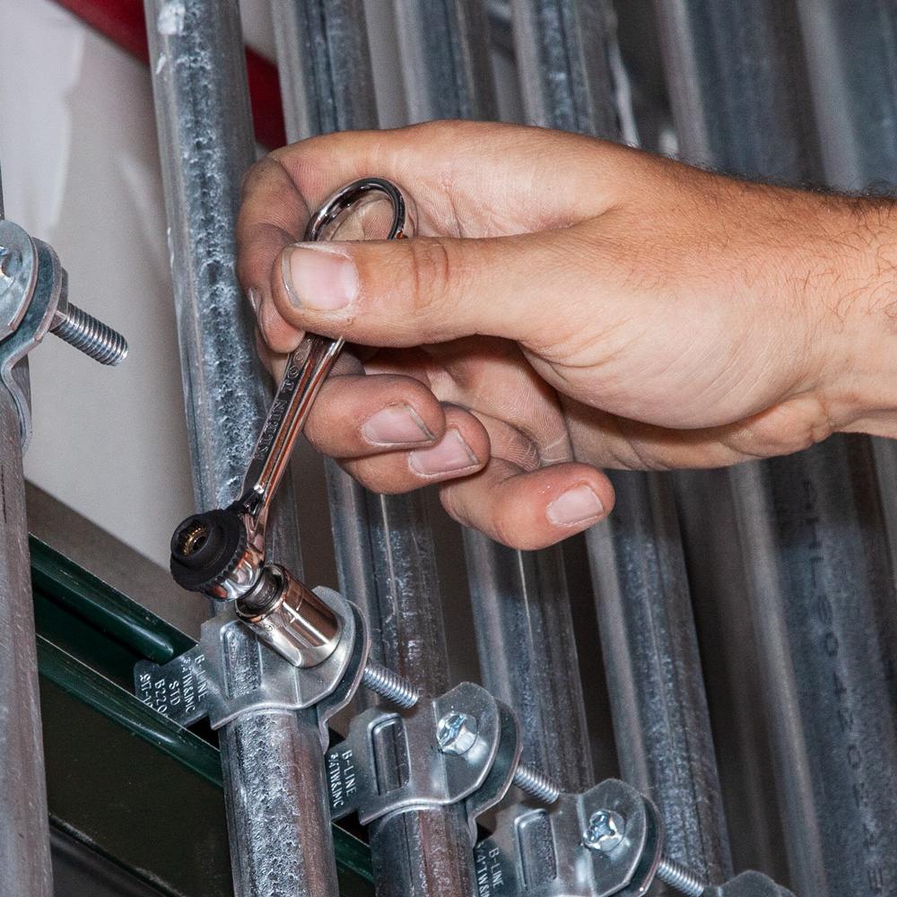 A hand holds the Klein 65200 mini ratchet by its finger loop while fastening a bolt on a metal conduit clamp.