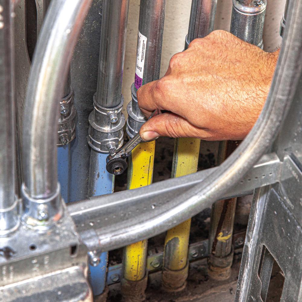 A worker's hand uses the slim-profile Klein mini ratchet to work on a fastener in a confined space between multiple pipes and conduits.