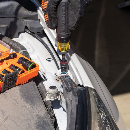 Action shot of a mechanic using the Klein Tools ratcheting screwdriver with a Flip-16 socket to remove a bolt from an automotive engine bay. The image demonstrates the tool's high-precision performance and ergonomic Cushion-Grip in a real-world application.