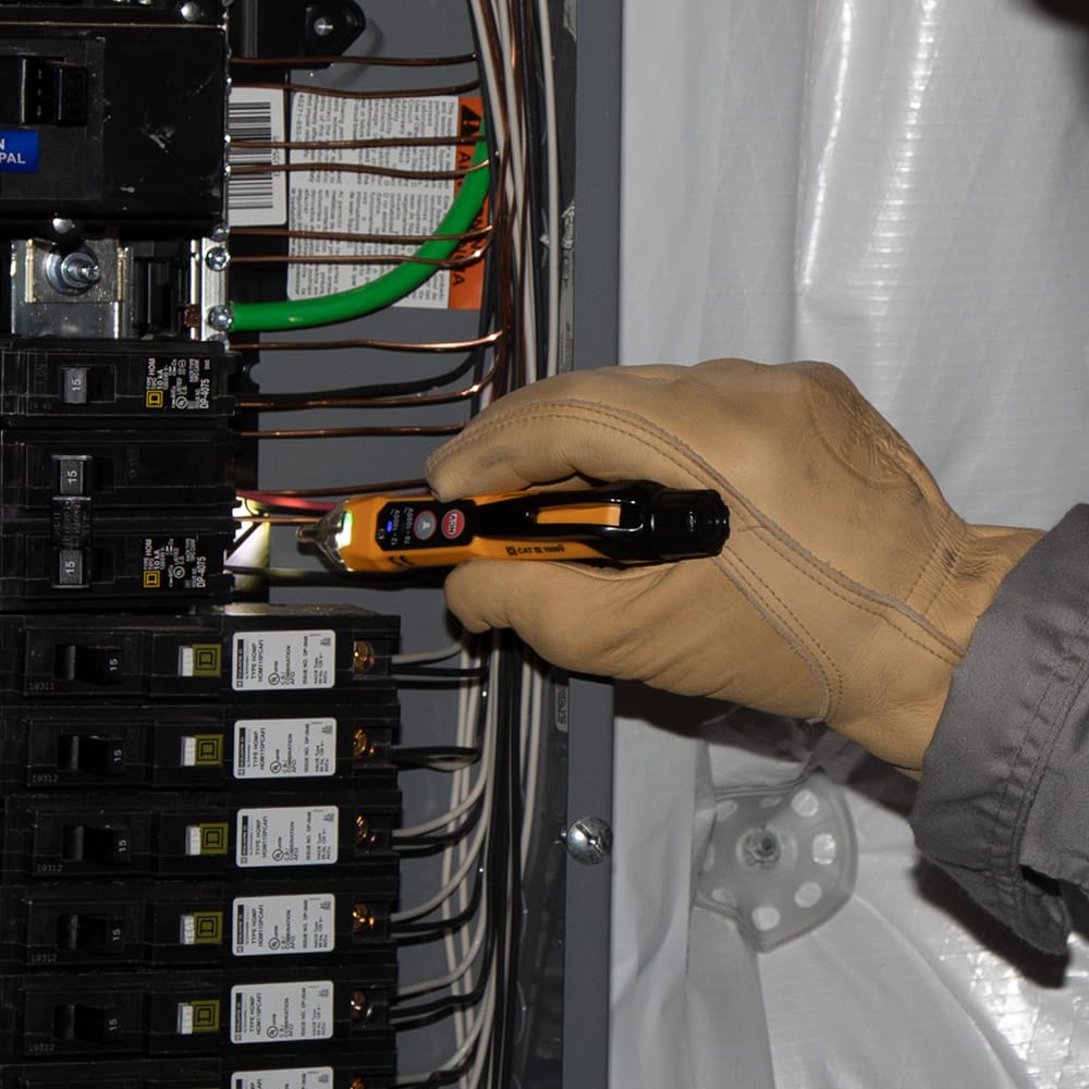 Action shot of an electrician wearing a protective leather glove using the NCVT3P non-contact voltage tester to inspect a circuit breaker panel. The integrated flashlight is active, illuminating the wires and demonstrating the tool's utility in dark electrical enclosures.