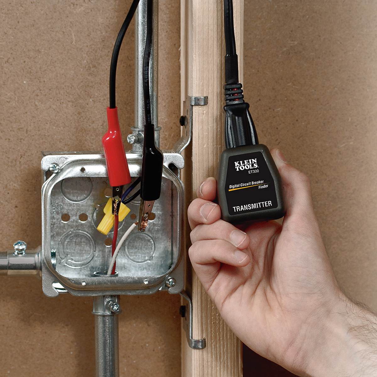 Action shot of a technician using the Klein Tools 69411 alligator clip adapter in an active electrical junction box. The red and black clips are shown connected to bare wire ends, feeding a signal to the Klein Tools ET300 Digital Circuit Breaker Finder transmitter held in the technician's hand.