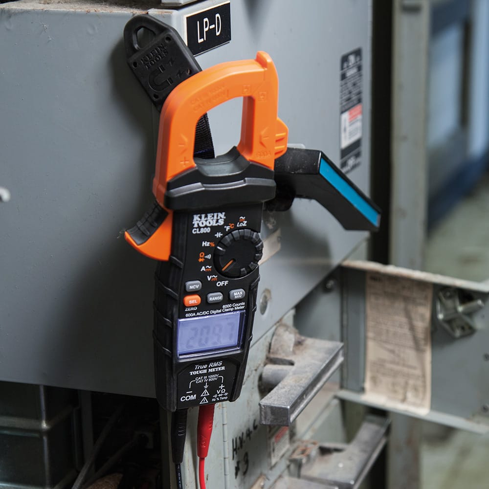 Action shot of a Klein Tools CL800 clamp meter mounted to a grey industrial electrical panel using the 69417 rare-earth magnetic hanger. The meter is held securely at eye level, demonstrating how electricians can easily monitor real-time data while troubleshooting complex circuits.