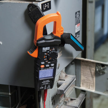 Action shot of a Klein Tools CL800 clamp meter mounted to a grey industrial electrical panel using the 69417 rare-earth magnetic hanger. The meter is held securely at eye level, demonstrating how electricians can easily monitor real-time data while troubleshooting complex circuits.