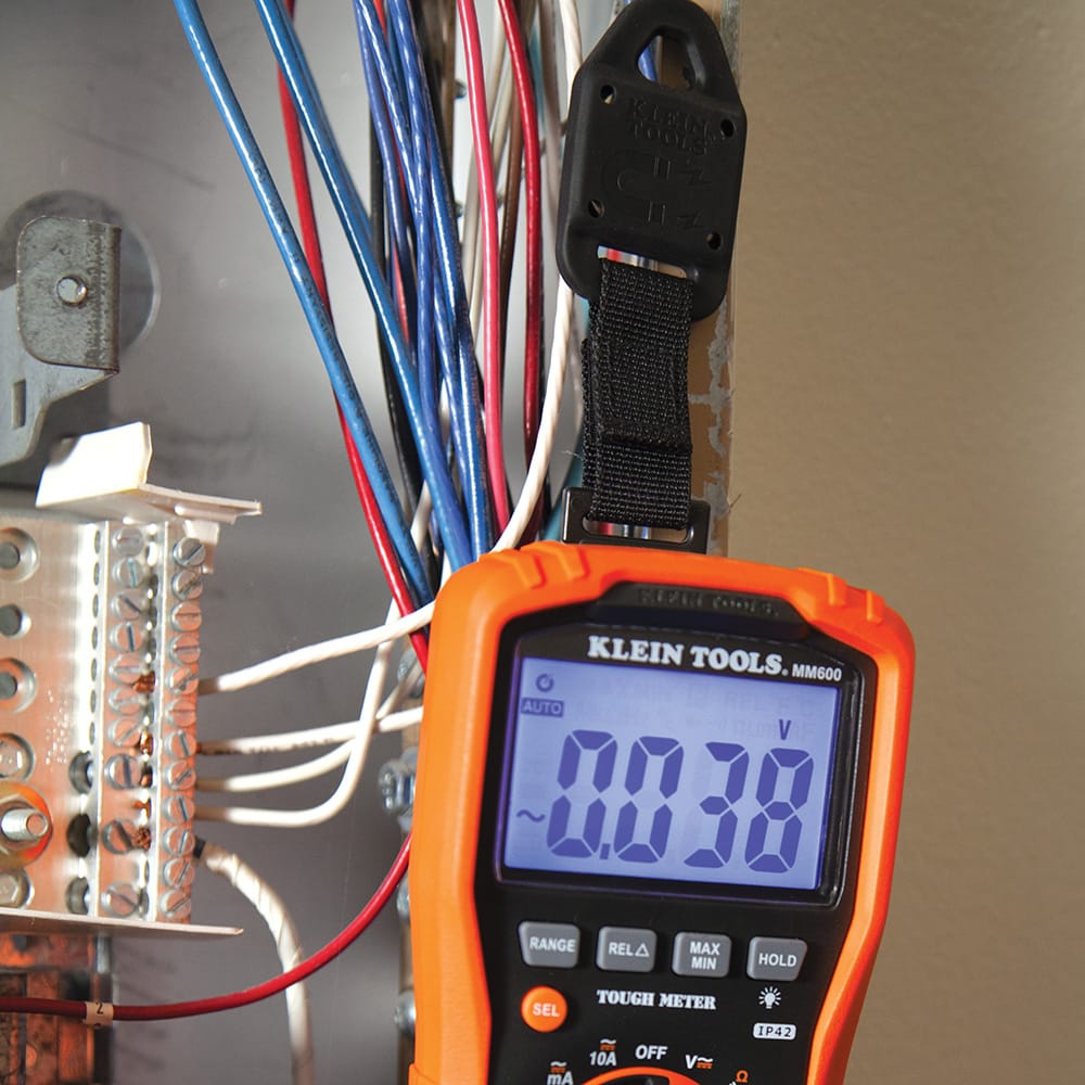 Action shot of the Klein Tools MM600 digital multimeter suspended from a steel enclosure using the magnetic strap hanger. The device's backlit display is clearly visible while the technician works within the wiring cabinet, highlighting the safety and convenience of hands-free operation.