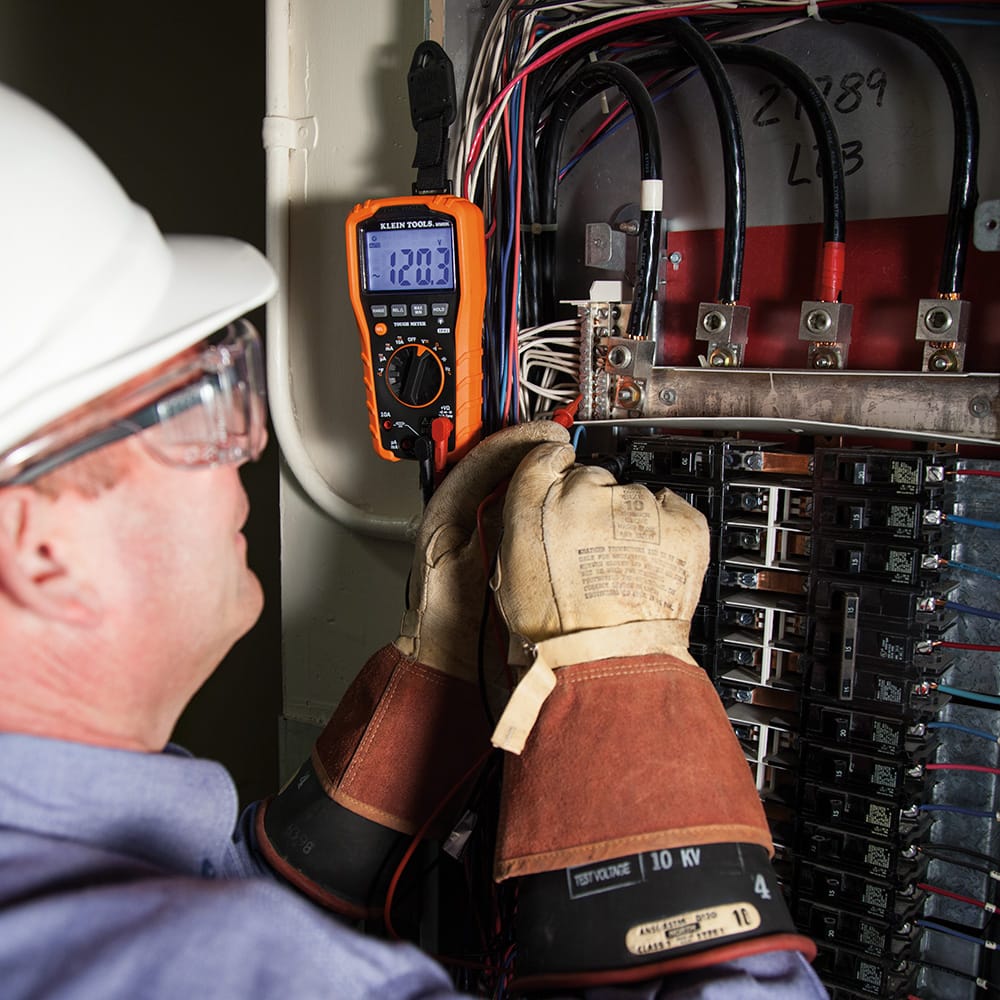 A professional electrician wearing protective gear and high-voltage gloves troubleshooting a main breaker panel. The Klein Tools multimeter is mounted to the side of the panel using the 69417 magnetic hanger, allowing the technician to use both hands for probing and safe manipulation of electrical components.