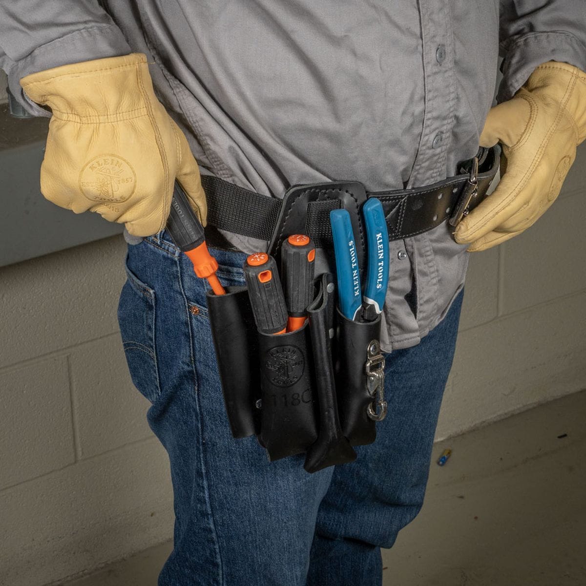 An electrician on a commercial jobsite wearing thick tan leather gloves reaches to pull a high-torque cushion-grip tool from the Klein Tools 3-piece 1000V insulated screwdriver set organized within a black leather tool belt pouch alongside blue-handled wire strippers while maintaining code compliance near electrical panels.