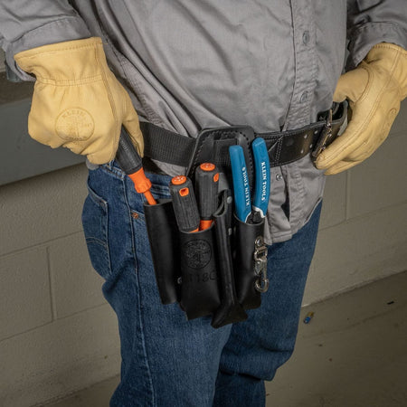 An electrician on a commercial jobsite wearing thick tan leather gloves reaches to pull a high-torque cushion-grip tool from the Klein Tools 3-piece 1000V insulated screwdriver set organized within a black leather tool belt pouch alongside blue-handled wire strippers while maintaining code compliance near electrical panels.