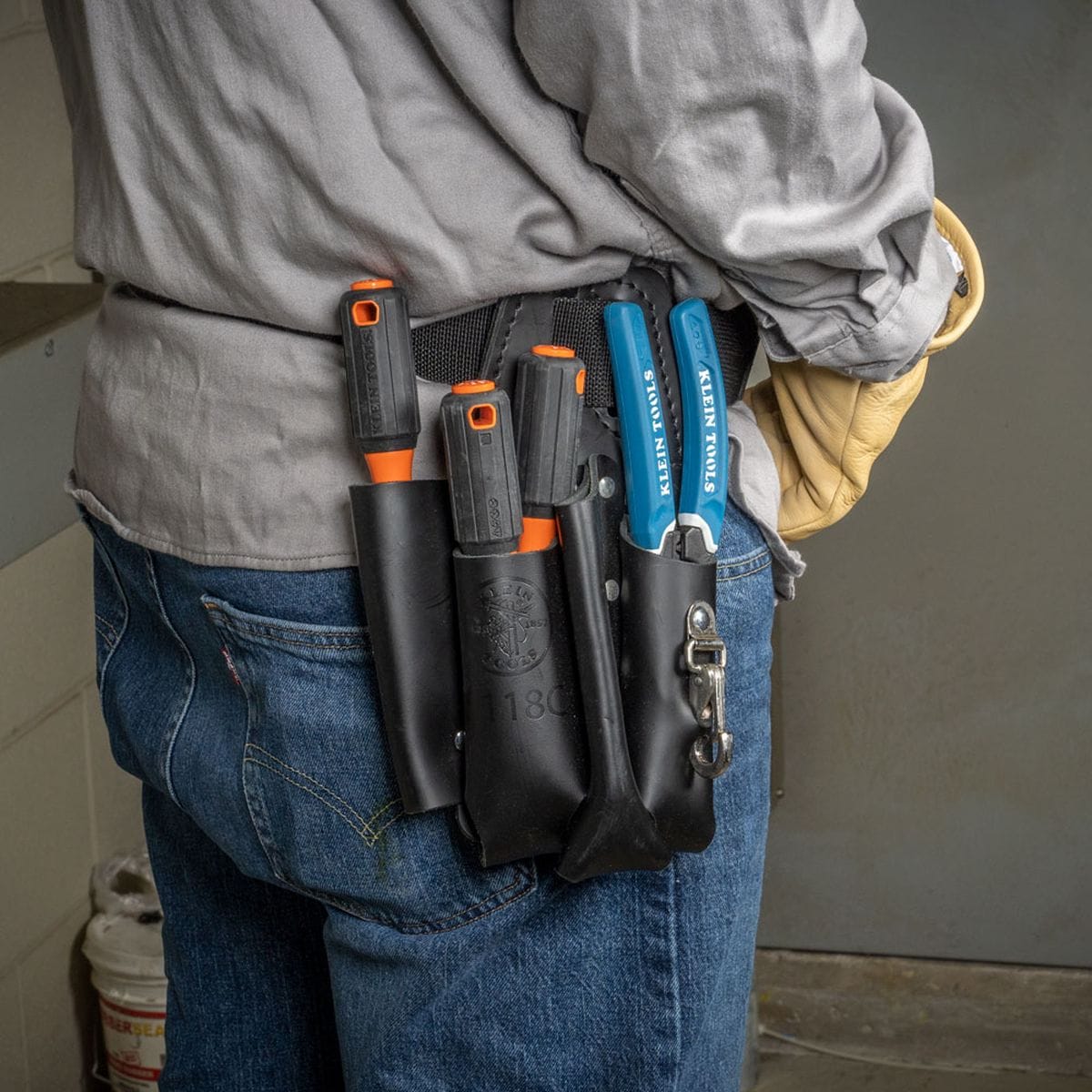 Side angle view of a trade professional wearing tan safety gloves and blue jeans displaying a black leather tool pouch attached to a work belt, housing multiple black and orange Klein Tools 1000V insulated drivers alongside blue high-dielectric wire cutting pliers for convenient hands-free tool storage.