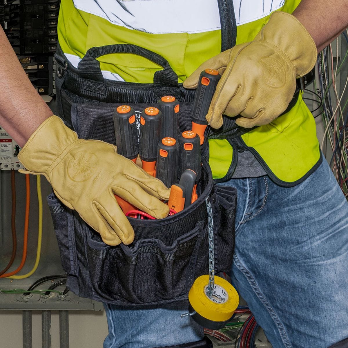 An electrician on a commercial site wearing thick tan leather gloves reaches to select a high-torque cushion-grip tool from the Klein Tools 6-piece 1000V insulated screwdriver set organized cleanly within a black canvas tool belt pouch alongside a yellow electrical tape dispenser reel while maintaining code compliance near electrical panel wiring runs.
