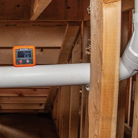 The angle gauge resting on top of a white PVC pipe running horizontally through wooden studs, displaying a reading of 2.0 degrees.