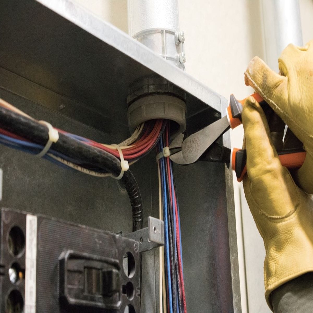 A close-up action view of a technician wearing tan leather work gloves utilizing the high-leverage insulated diagonal cutting pliers to cleanly snap through a plastic cable tie surrounding a high-voltage wire bundle inside a steel service box.