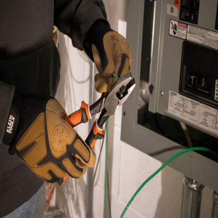 A worker wearing padded mechanical gloves holding the high-leverage insulated side-cutting linesman pliers from the 94130 kit to carefully strip and prepare a green insulated copper conductor wire next to an open circuit breaker panel.