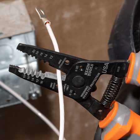 A close-up view of a black oxide Klein Tools insulated wire stripper cutter tool being used to strip insulation off a solid white electrical cable wire, showcasing the integrated size gauge slots labeled for solid and stranded AWG conductor parameters.