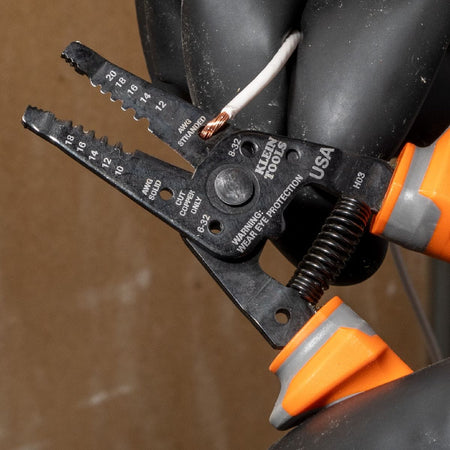 An electrician wearing industrial black rubber work gloves holds a solid copper conductor inside the jaw alignment notches of a Klein Tools 1000V rated insulated wire stripper tool, preparing to perform a clean insulation strip cycle near a terminal housing unit.