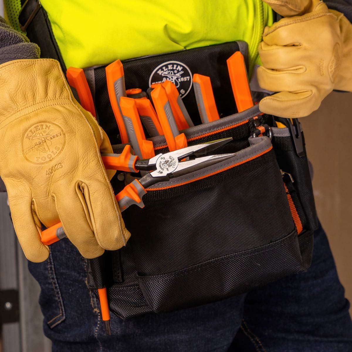 A close-up field view of an electrician wearing branded tan leather safety work gloves and a high-visibility yellow vest, organizing a set of matching orange and gray Klein Tools 1000V insulated pliers and wire strippers cleanly inside a black canvas tool belt pouch.