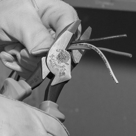 A close-up action view of an operator utilizing a pair of heavy-duty Klein Tools 2139NERINS insulated side-cutting lineman pliers to cleanly slice through a bundle of multi-conductor electrical wires, demonstrating the cutting force of the induction-hardened blades.