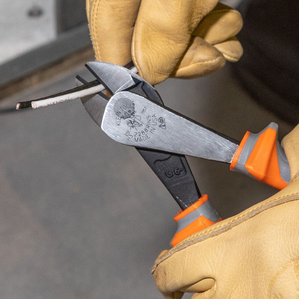 An electrician wearing heavy tan leather work gloves uses a pair of Klein Tools 2288RINS insulated diagonal cutters to clip a multi-conductor electrical wire core, demonstrating the heavy leverage cutting jaw action and non-slip protective orange and gray thumb guard handle profile.